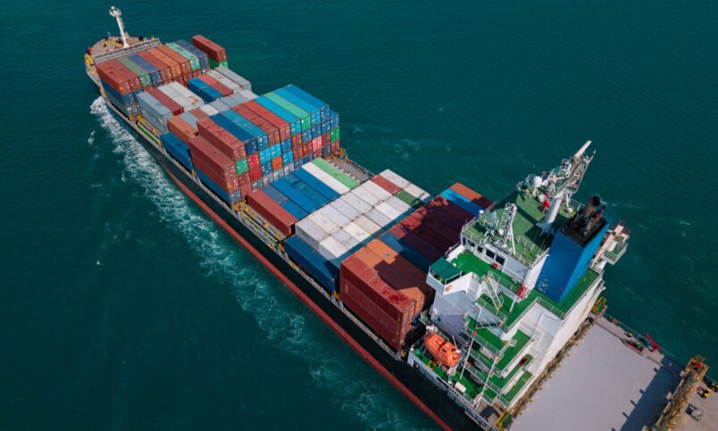 A massive container ship sails across calm blue waters, carrying colorful shipping containers, representing global trade and logistics.